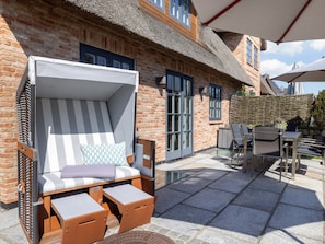 Eine Terrasse mit einem Gebäude mit Reetdach, Sitzgelegenheiten mit Kissen und einem Tisch mit Stühlen.
