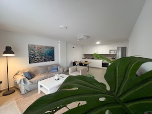 A modern living room with a sofa, coffee table, and a painting. There is a kitchen area with cabinets and appliances in the background. - Vacation Apartment 'Sotavento Suites' with Sea View, Shared Pool and Wi-Fi (El Médano)