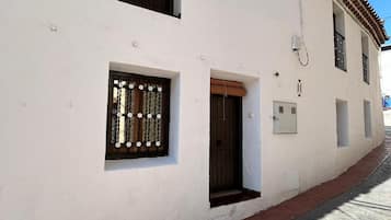 Un edificio blanco con techo de tejas, una ventana pequeña con una reja metálica y una puerta con marco marrón.