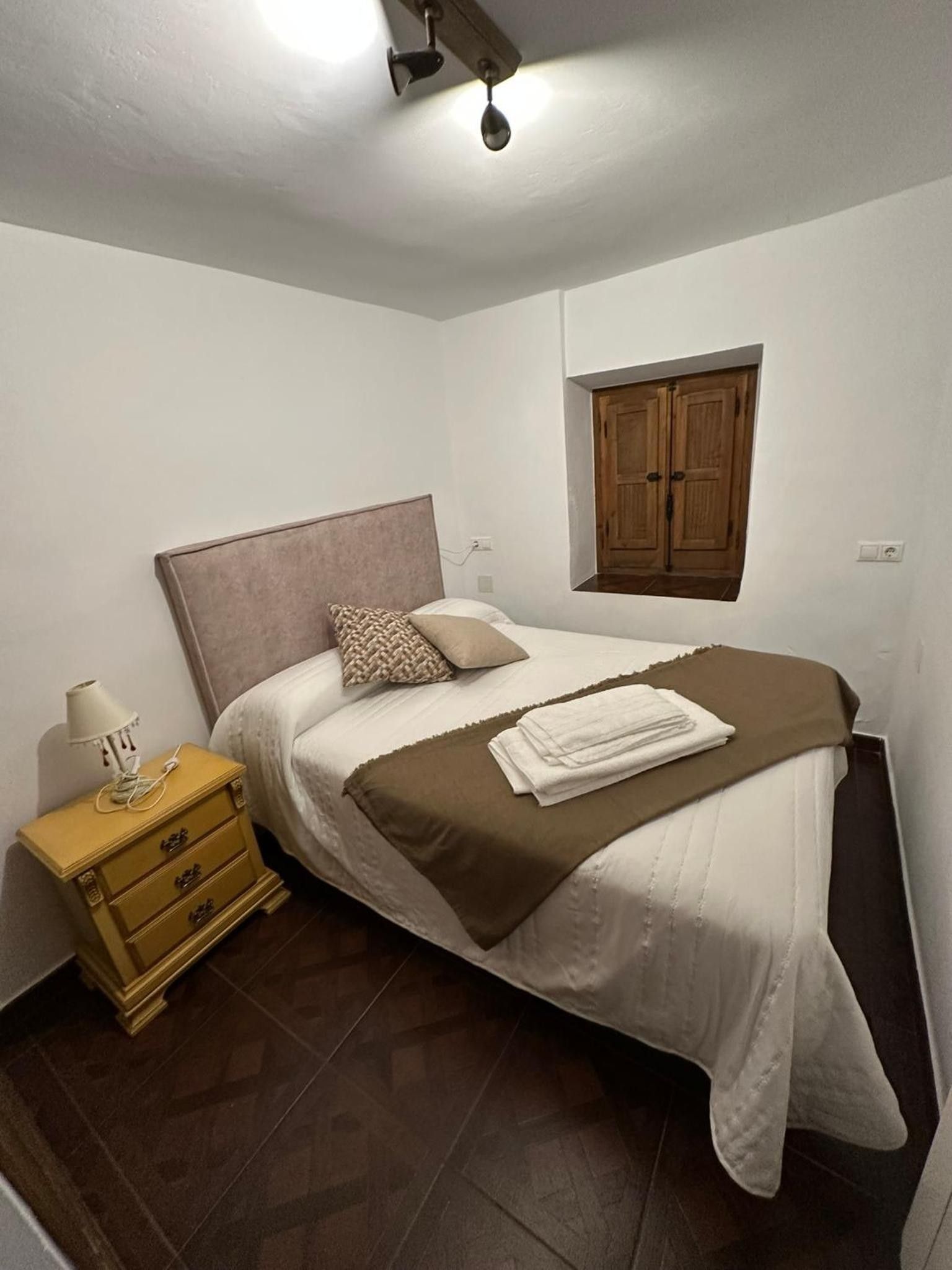 Un dormitorio con cama, mesita de noche, lámpara, ventana con persiana de madera y una lámpara de pared.