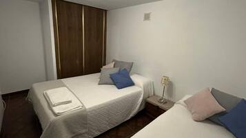 Habitación de hotel con dos camas, un armario de madera, una mesita de noche con lámpara y un aplique de techo.