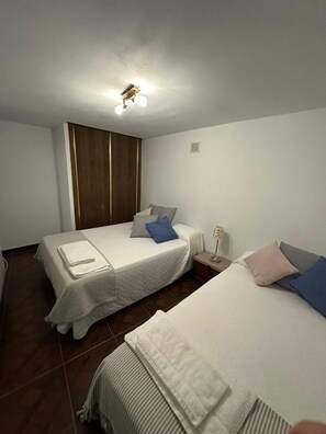 A hotel room with two beds, a wooden wardrobe, a bedside table with a lamp, and a ceiling light fixture.