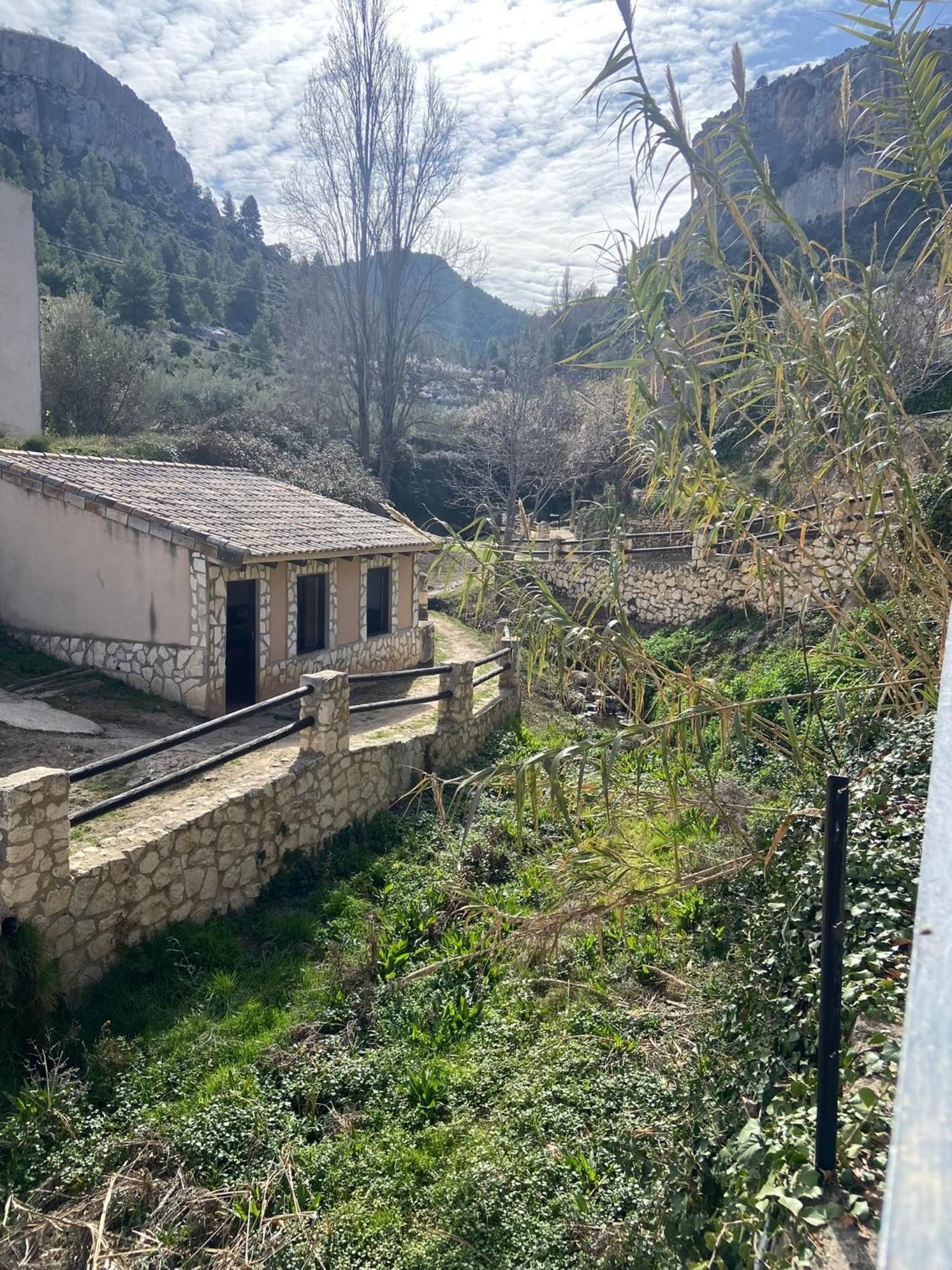 Un edificio de piedra con techo de tejas, rodeado de vegetación y montañas al fondo.
