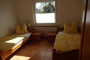 Ein kleines Zimmer mit zwei Einzelbetten, jeweils mit Kissen und Nachttisch. Über den Betten befindet sich ein Fenster und darunter ein Heizkörper.