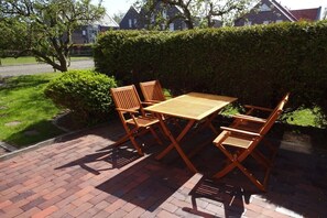 Une terrasse avec une table en bois et quatre chaises, entourée d’une haie et bordée d’un chemin en briques.
