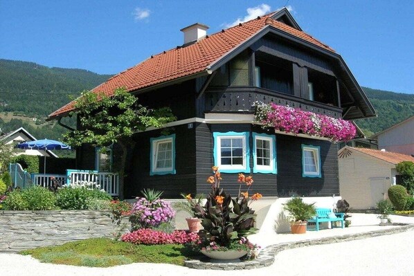 A two-story house with a red-tiled roof, a balcony with flowers, and a stone wall in the foreground. - Haus Thon holiday apartment garden view (Bodensdorf)