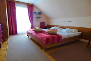 A hotel room with a bed, a wooden headboard, a nightstand, a desk, a chair, a bookshelf, and a window with curtains. - Haus Thon holiday apartment garden view (Bodensdorf)