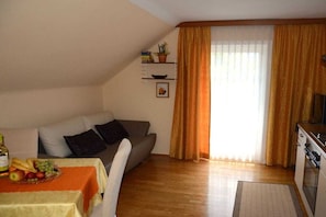 A living room with a sofa, a dining table with fruit, a kitchen area, and a window with curtains. - Haus Thon holiday apartment garden view (Bodensdorf)