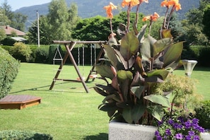 A garden with a swing set, a concrete planter with large leaves, and a wooden deck. - Haus Thon holiday apartment garden view (Bodensdorf)