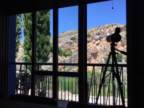 A telescope on a tripod outside a window, with a view of rocky terrain and trees.