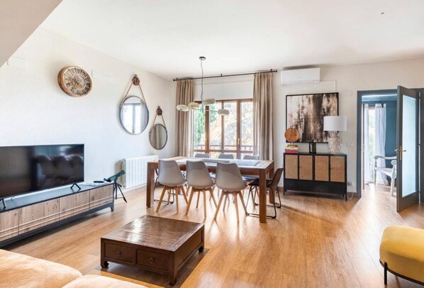 A modern living room with a flat-screen TV, wooden dining table, and chairs.