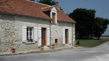 Une maison en pierre avec un toit recouvert de tuiles rouges, des volets blancs et un petit porche agrémenté de plantes en pot.
