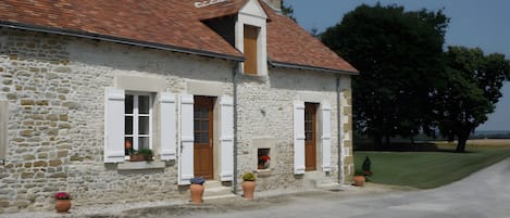 A stone house with a red-tiled roof, white shutters, and a small porch area with potted plants.