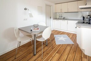 A compact kitchen with white cabinets, a dining table with chairs, a wooden floor, and a star-patterned rug. - "Tied" at Uns to Hus (Juist)