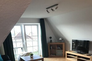 A living room with a television, wooden furniture, a bookshelf, and a large window with curtains. - Holiday apartment with 2 bedrooms (Hooksiel)