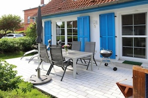 Eine Terrasse im Freien mit Tisch, Stühlen, einem Grill und einem blauen Haus mit Fensterläden.