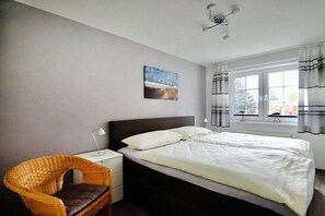 A bedroom with a bed, a nightstand, a chair, a window with curtains, and a painting on the wall. - Mühlenpark 1 W 1.2 "Crane Train" (Prerow)