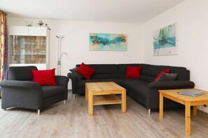 Ein Wohnzimmer mit einer dunkelgrauen Sofagarnitur, zwei Holztischen und roten Kissen.
