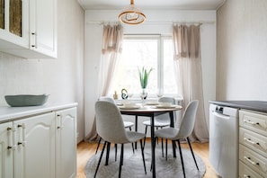 Een eetgedeelte met een ronde tafel, vier stoelen, een vaas met een plant en een raam met gordijnen.