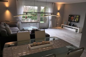A modern living room with a glass dining table, a sofa, a TV, and a window with curtains. - Holiday apartment with 1 bedroom (Norddeich)