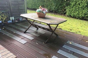Een houten terras met een tafel en stoelen, omgeven door een tuin en een haag.