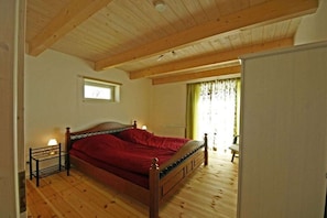 Ein Schlafzimmer mit einem Holzbett, einem Nachttisch mit Lampe, einem Fenster mit Vorhängen und Blick auf eine grüne Außenanlage.