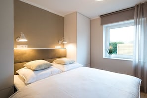 Ein Hotelzimmer mit einem großen Bett, zwei Nachttischlampen, einem Fenster mit Vorhängen und Blick ins Grüne.