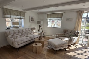 A living room with a sofa, armchair, and round coffee table. There are windows with curtains and a lamp on a tripod stand. - Holiday home in Vollerwiek with sauna (Vollerwiek)