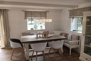 A dining area with a wooden table, white chairs, a bench with cushions, a hanging light fixture, and a window with curtains. - Holiday home in Vollerwiek with sauna (Vollerwiek)
