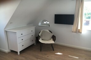 A room with a white dresser, a chair with a fluffy cushion, a mounted TV, and a window with curtains. - Holiday home in Vollerwiek with sauna (Vollerwiek)