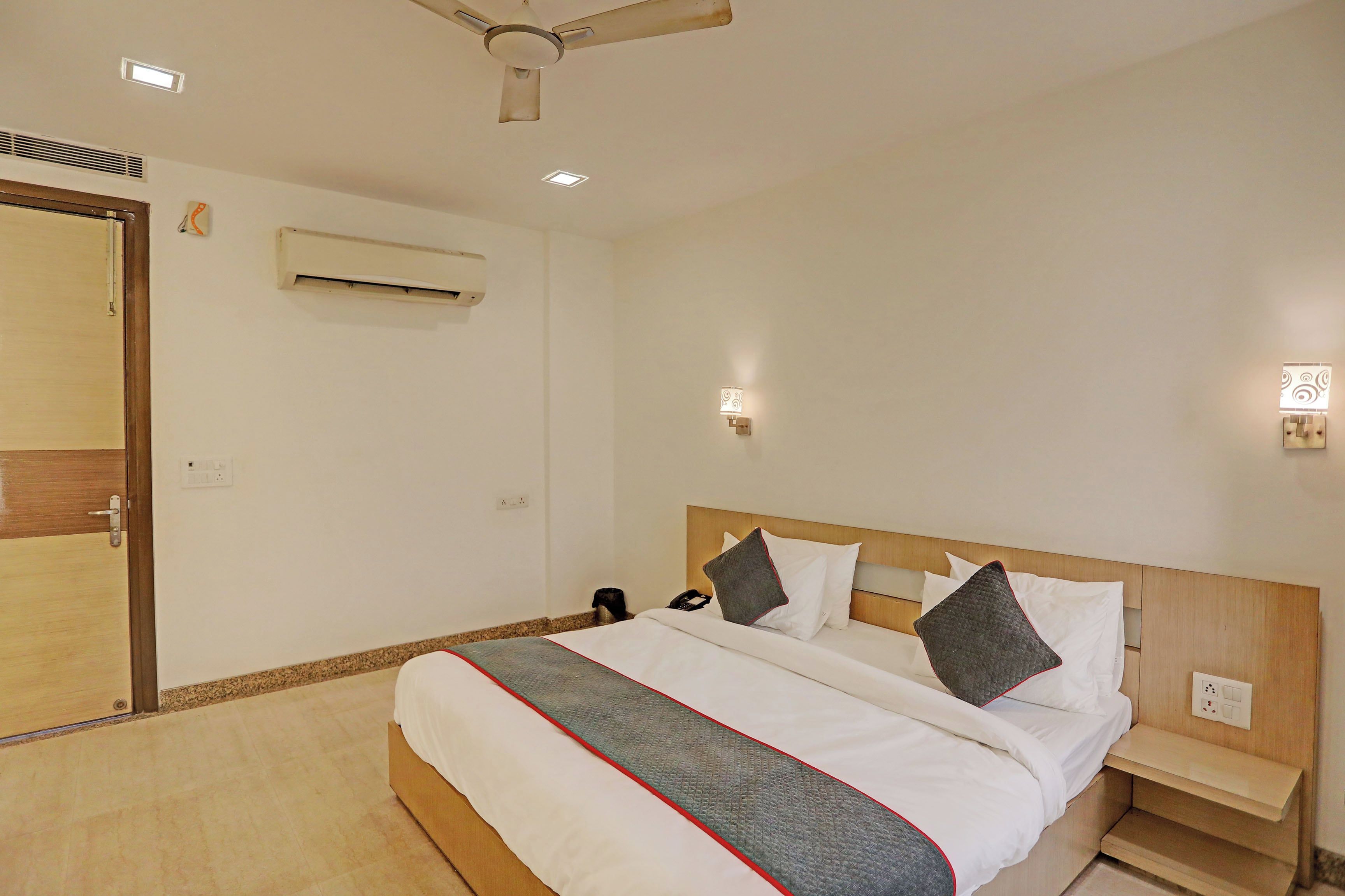A hotel room with a bed, bedside tables, a ceiling fan, an air conditioning unit, and wall-mounted lights.