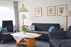 Ein modernes Wohnzimmer mit einem grauen Sofa, einem Holztisch und einem Sessel. Zwei Stehlampen, eine Vase mit Blumen und gerahmte Bilder schmücken die Wand.