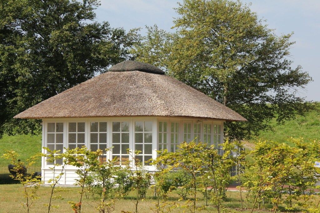 Une petite construction à toit de chaume, dotée de grandes fenêtres et entourée de verdure et d’arbres.