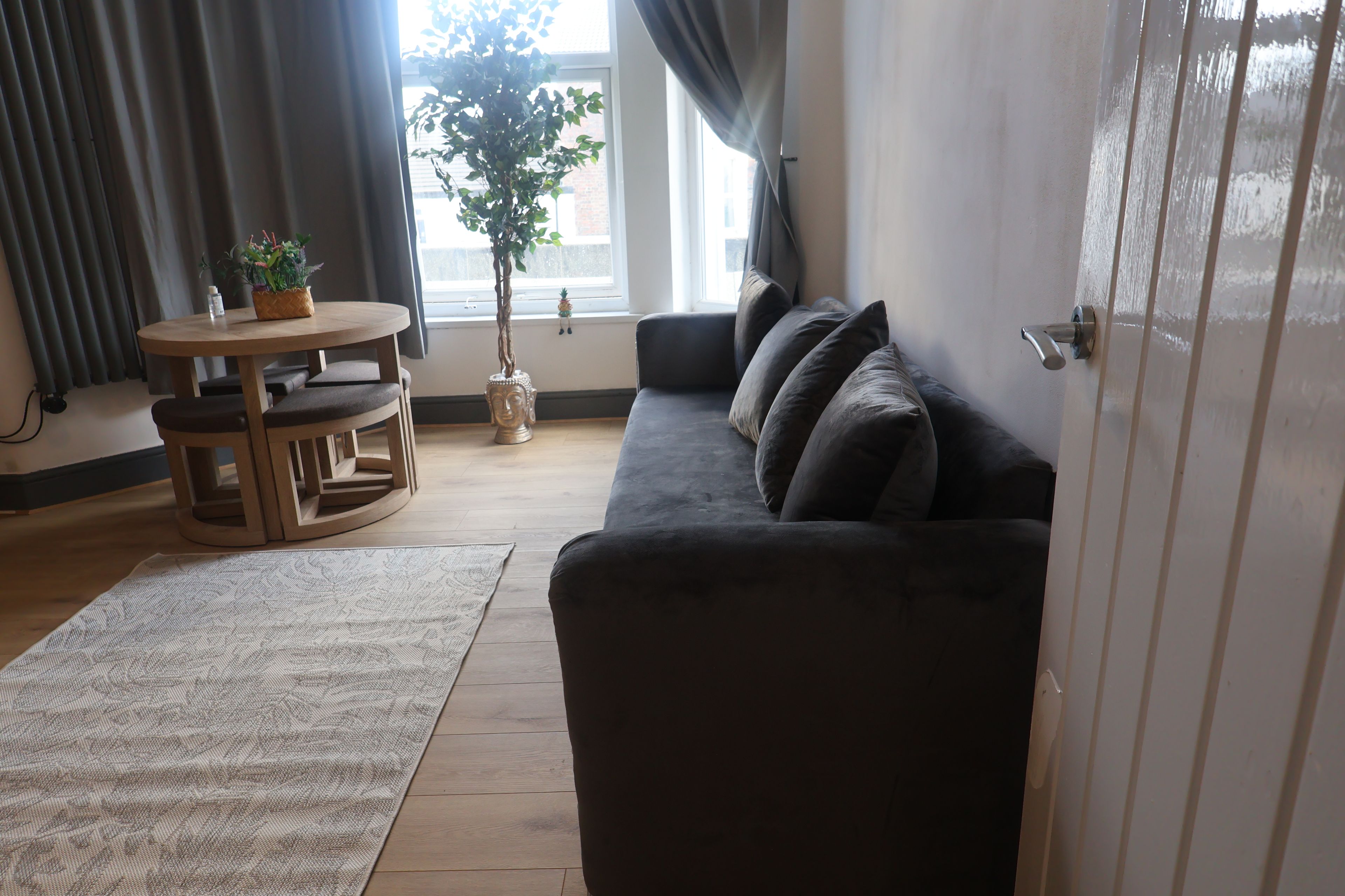 Un salon avec un canapé de couleur foncée, une table ronde en bois entourée de chaises, une plante en pot et une fenêtre avec des rideaux.