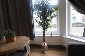 A room with a wooden table, a potted plant, a window with curtains, and a small Buddha statue.