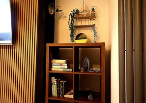 A bookshelf with books and decorative items, a wall-mounted shelf with plants, and a window with blinds.
