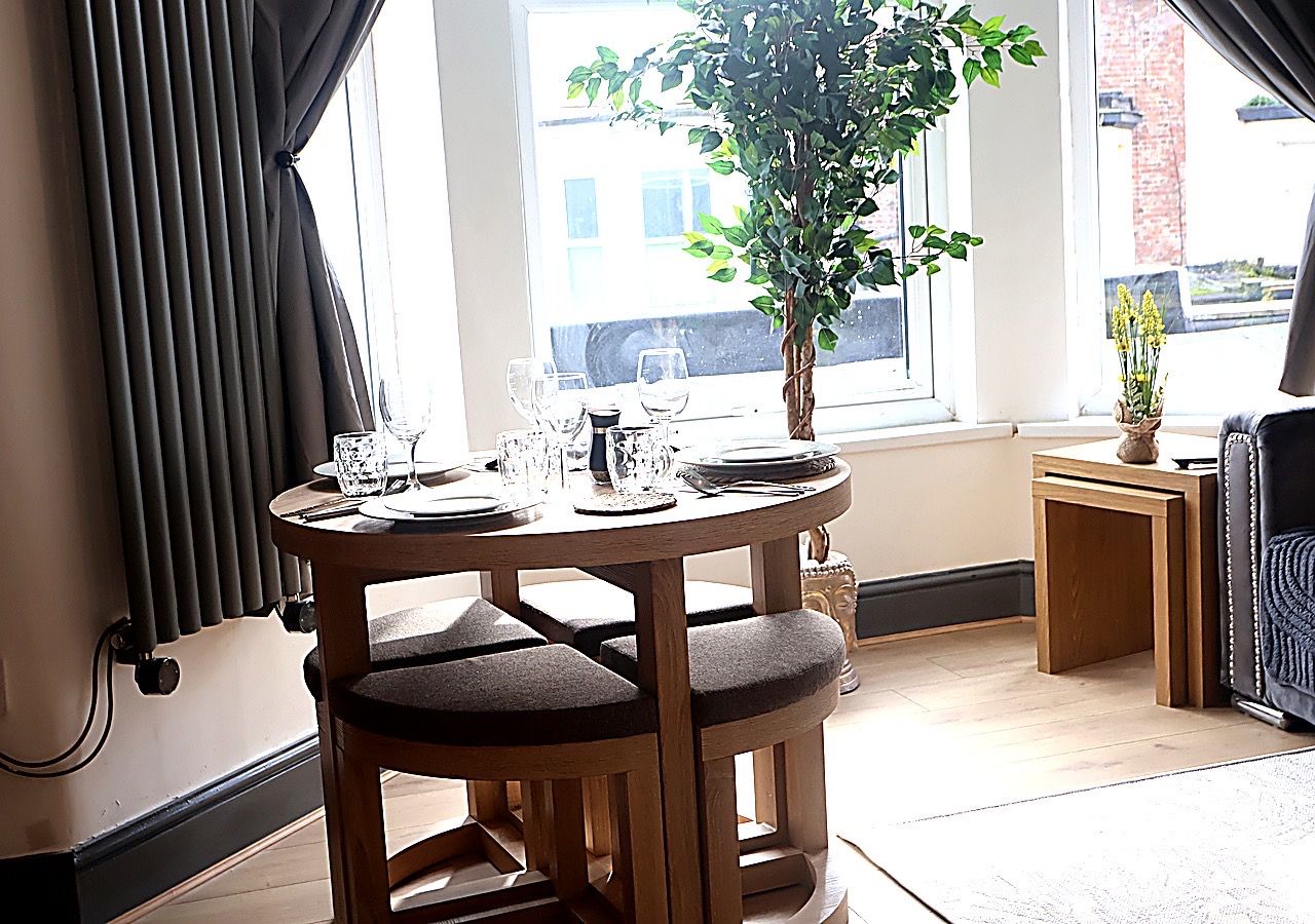Une table ronde en bois, entourée de quatre chaises, avec une plante en pot et une fenêtre donnant sur des immeubles.