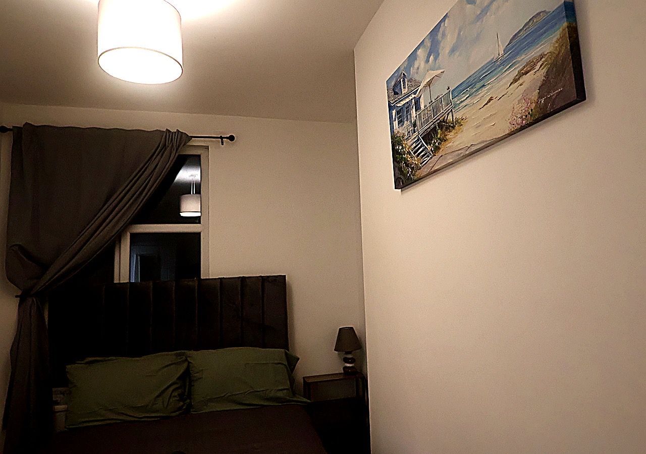 A bedroom with a bed, a nightstand, a lamp, a painting on the wall, and a window with curtains.