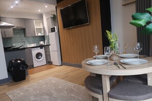 A modern kitchen with a built-in washing machine, a dining area with a round table set for four, and a flat-screen TV mounted on the wall.