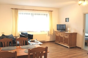 A living room with a wooden dining table, chairs, a television on a wooden cabinet, and a window with curtains. - Baltic Sea dike (07) (Ostseebad Prerow)
