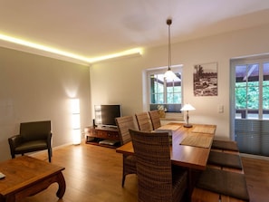 A modern living room with a wooden dining table, comfortable seating, a flat-screen TV, and a large window with a view of greenery. - Holiday Apartment With 3 Bedrooms (Braunlage)