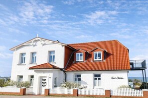 A two-story house with a white facade and red-tiled roof, featuring a white picket fence and a small balcony. - Holiday apartment with 1 bedroom (Wittdün)