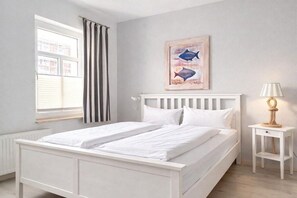 A bedroom with a white bed, a window with curtains, a bedside lamp, and a painting on the wall. - Holiday apartment with 1 bedroom (Wittdün)