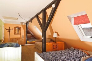 Ein Schlafzimmer mit geneigter Decke, einem Bett mit gestreifter Bettwäsche, einem Holzschrank, einem Spiegel und einem Fenster mit rotem Rollo.