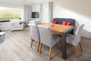 Ein modernes Hotelzimmer mit einem Holztisch, grau gepolsterten Stühlen, einem grauen Sofa, einem Fernseher und einem Fenster mit Vorhängen.