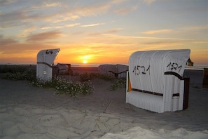 Zwei weiße Korbstühle mit den Nummern 64 und 4524, platziert an einem sandigen Strand mit Sonnenuntergang im Hintergrund.
