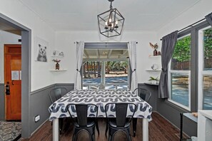 A dining area with a table covered in a patterned tablecloth, surrounded by chairs. A hexagonal chandelier hangs from the ceiling, and a window with curtains is visible. - PET FRIENDLY Boulder Bay Retreat with HOT TUB Under the Pines, Remodeled Kitchen, Multiple Decks, Outdoor Games & Cozy Mountain Charm! (Big Bear Lake)