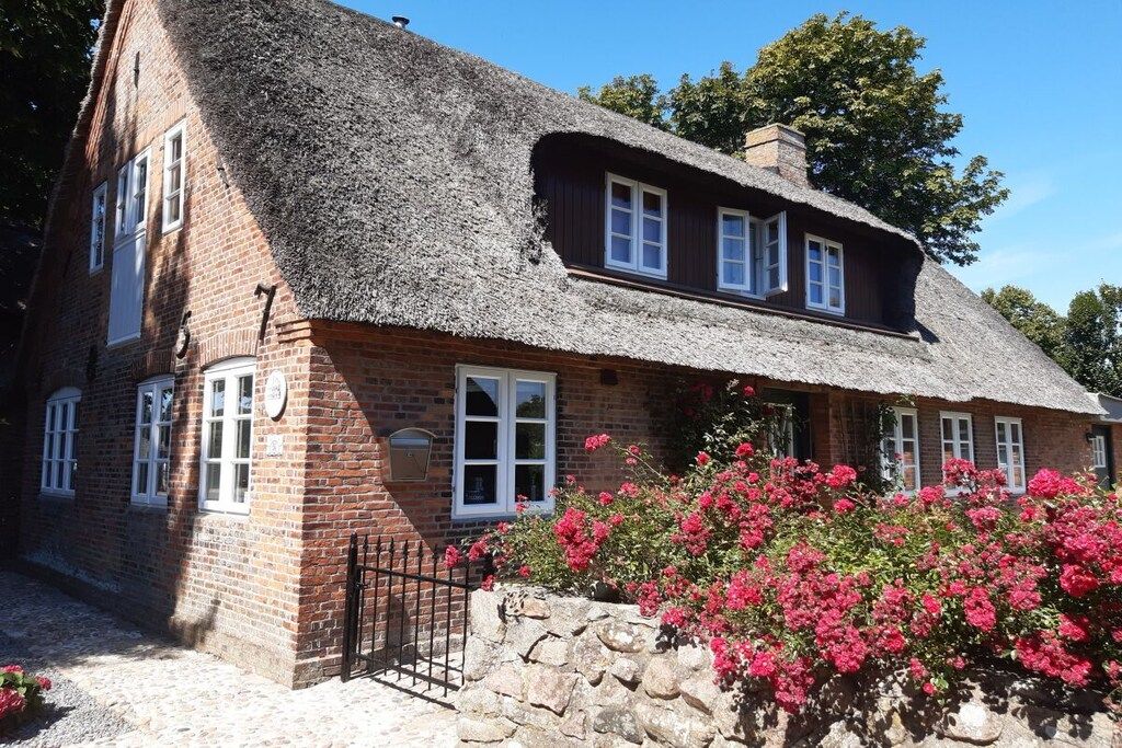 A traditional brick house with a thatched roof, a stone wall, and a garden with pink flowers.