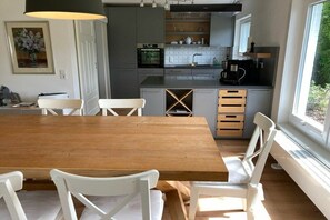 A modern kitchen with a wooden dining table and white chairs, a built-in oven, and a window providing natural light. - Holiday home in Nebel with swimming pool (Nebel)