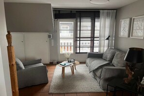 Ein Wohnzimmer mit grauem Sofa, einem Couchtisch und einer Lampe. Im Hintergrund befindet sich ein Fenster mit Jalousien und eine Tür.